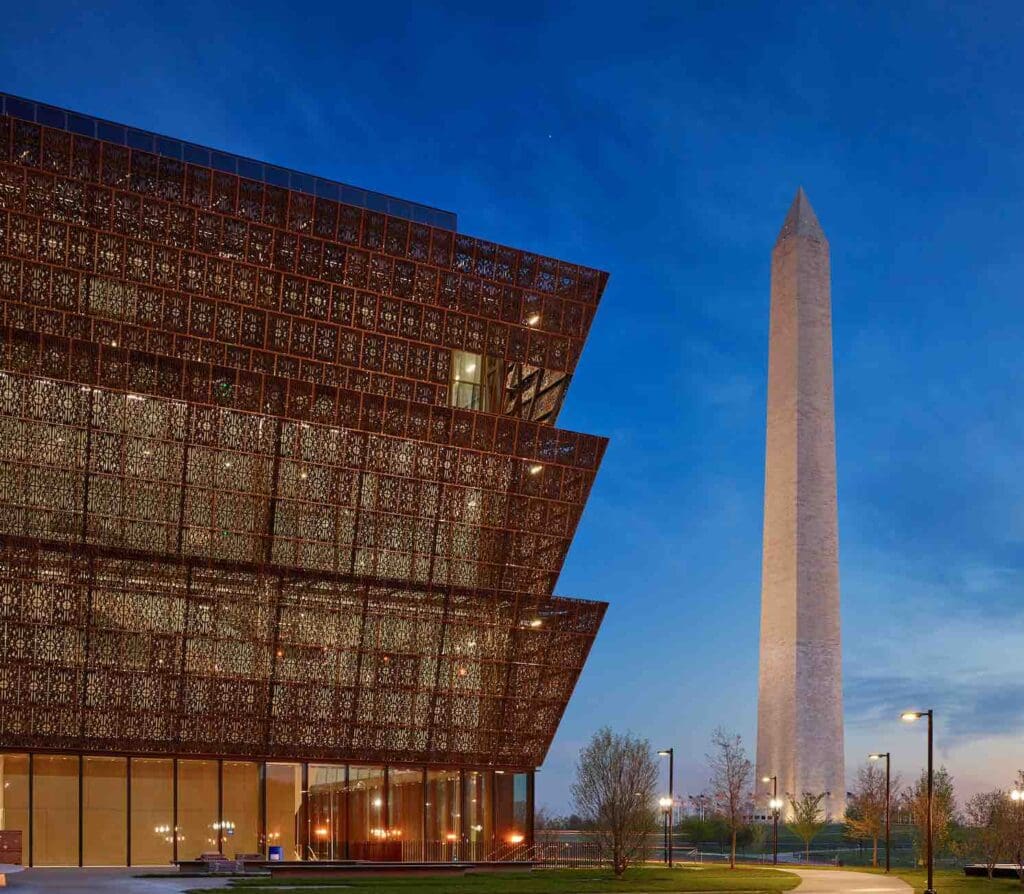 Modern building with lattice exterior panels, Washington Monument in background at twilight, lit landscape