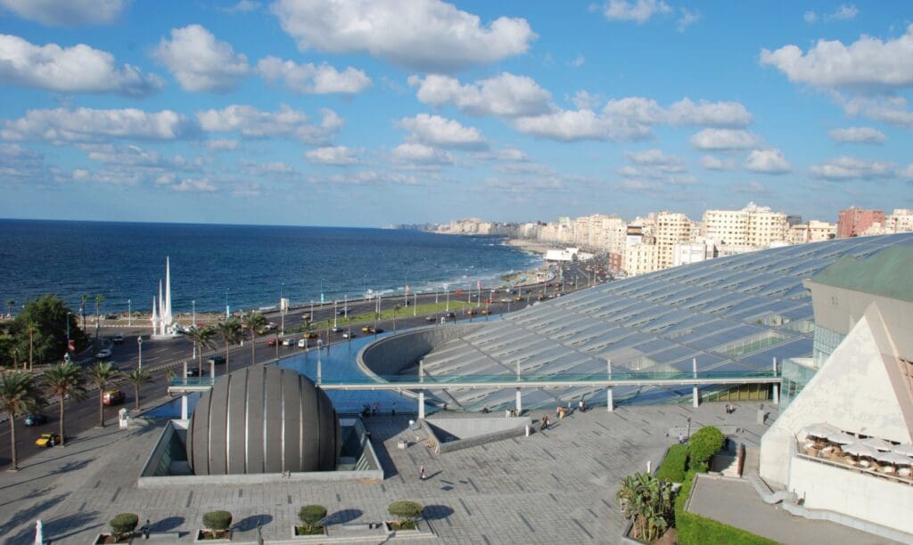 Coastal urban landscape with a modern structure, palm-lined road, ocean view, buildings, and clear sky.