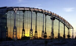 Large glass building with curved roof and steel trusses, reflecting sunset rays, clear sky background