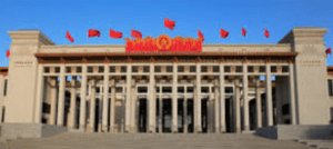 Large building with grand facade, tall columns, red flags, raised platform, and central emblem.