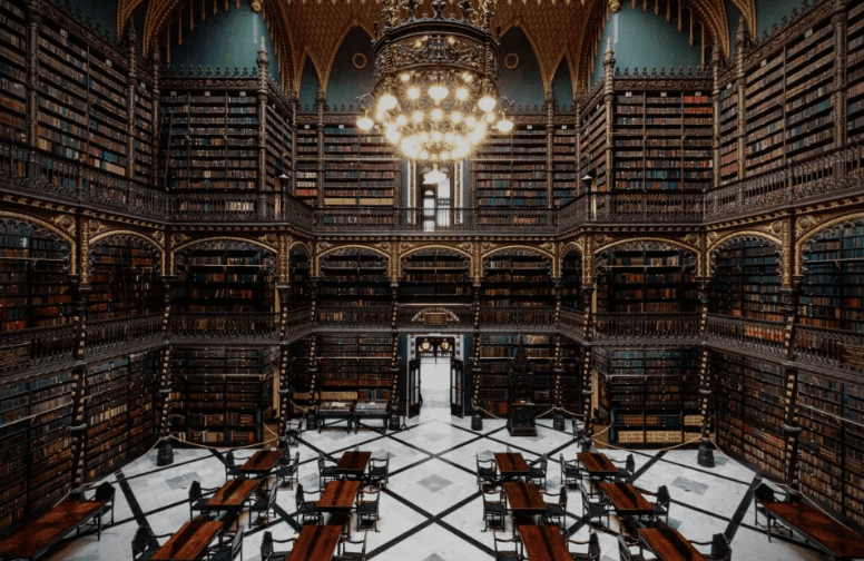 Grand library with ornate bookshelves, chandelier, and wooden tables on tiled floor.