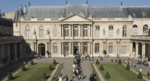 Historic building with classical columns, arched windows, sculptures, and landscaped courtyard with people