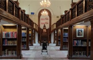 Library with wooden shelves, central aisle, balconies, chandeliers, India anniversary banner, statue.