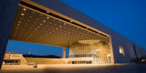 Modern architectural structure with a lit canopy roof over a glass-fronted building at twilight.