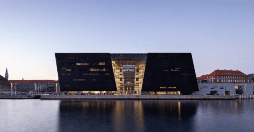Modern building with black facade, large glass central section, beside a body of water at twilight.