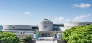 Modern building with cylindrical structures, central entrance, and surrounding trees on a clear day.