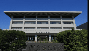 Modern building with rectangular windows, stone sign with inscriptions, shrubs in front, clear sky