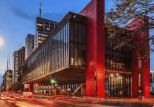 Elevated modern building with red pillars and glass panels in an urban area, captured at dusk.