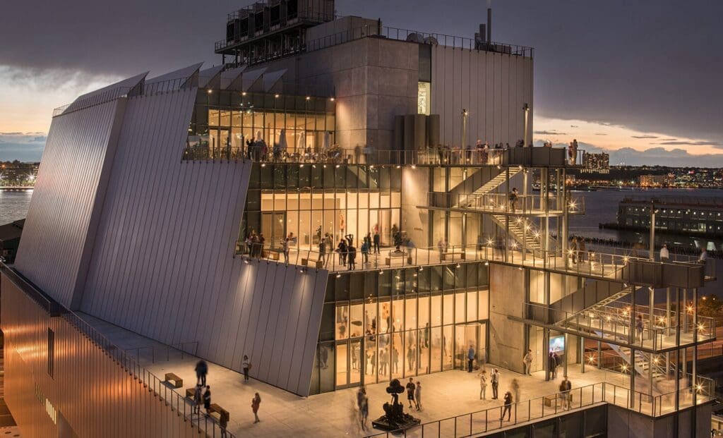 Modern multi-level building with glass windows overlooking a waterfront, cityscape in background.