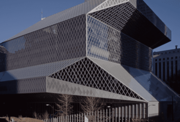 Modern angular building with geometric glass-and-steel exterior, surrounded by trees and urban structures.