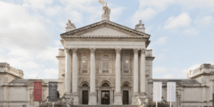 Neoclassical building with columns, pediment, statues, large windows, entrance doors, and display banners.