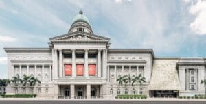 Large neoclassical building with a dome, tall columns, palm trees, modern annex, and banners.
