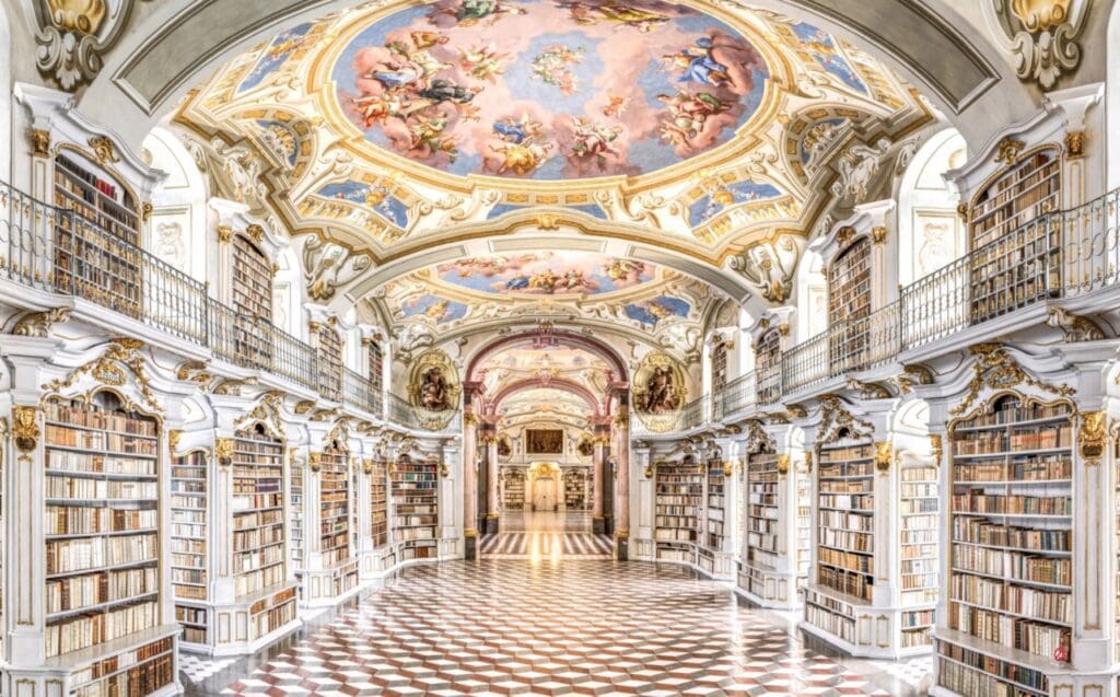 Ornate library interior with frescoed ceiling, gold detailing, bookshelves, and checkered floor.