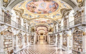 Ornate library interior with frescoed ceiling, gold detailing, bookshelves, and checkered floor.