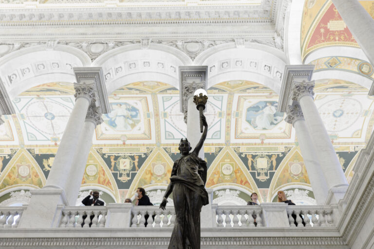 Grand hall with high frescoed ceiling, large columns, a statue with a torch, and people on a balcony