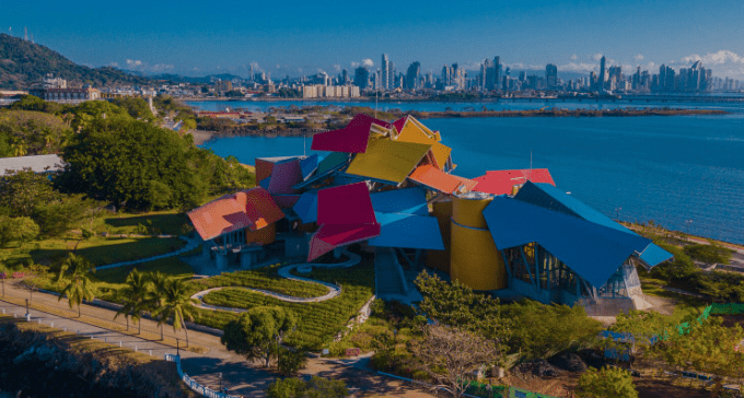 Aerial view of a multicolored roof building by water, with city skyline and greenery in the background.