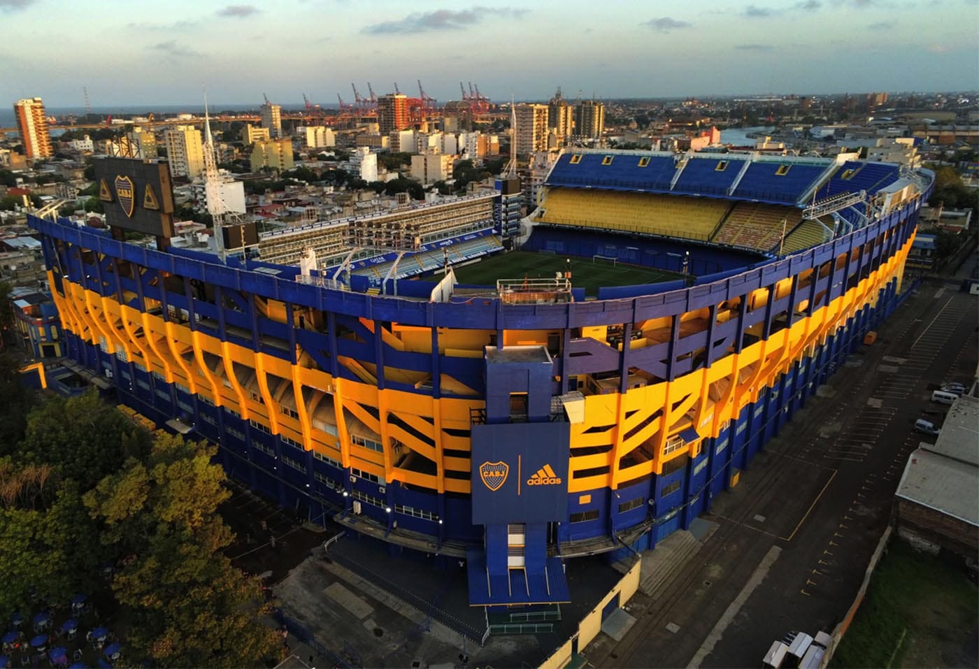 Aerial view of a blue and yellow sports stadium surrounded by an urban cityscape and port.