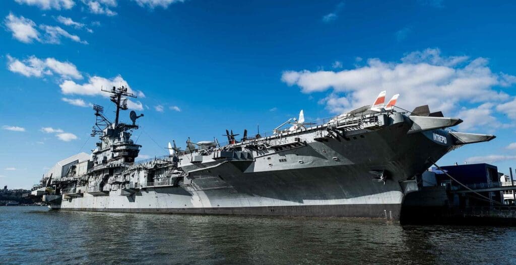 Aircraft carrier docked at harbor under blue sky with scattered clouds, equipped with military gear.