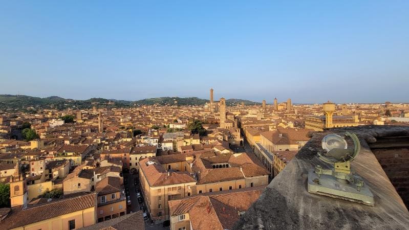 Panoramic view of an ancient city with terracotta rooftops, towers, and hills under a clear sky.