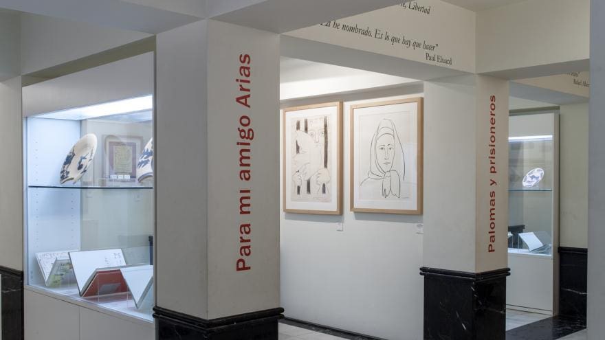 Art exhibition with framed drawings, ceramic pieces in glass cases, and pillars featuring Spanish text.