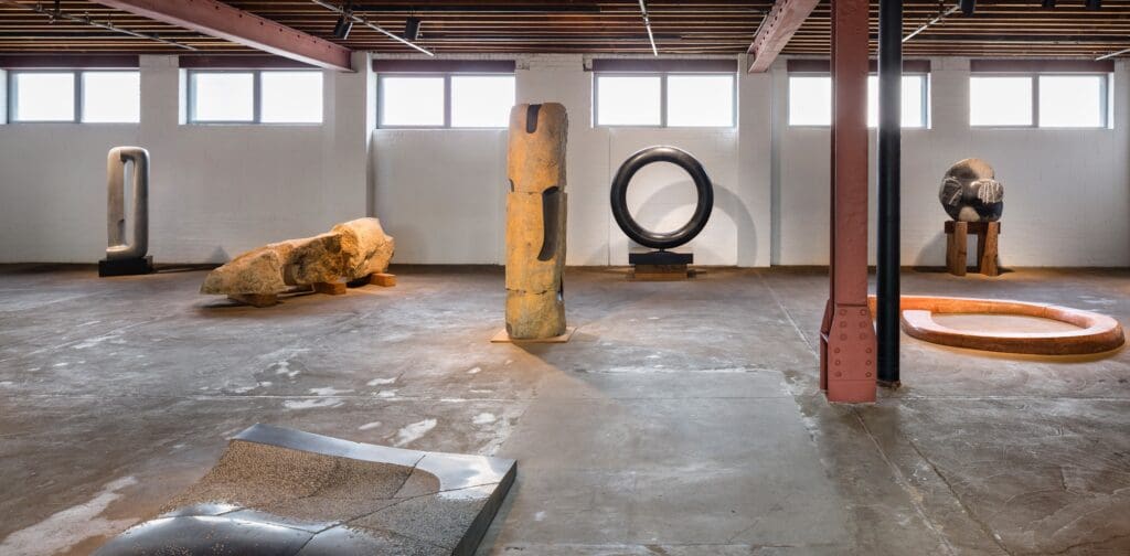 Art gallery with large abstract stone and metal sculptures, concrete floor, exposed beams, and natural light.