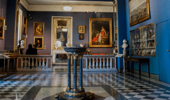 Art gallery with checkered floor, blue walls, framed paintings, sculptures, and an ornate basin.