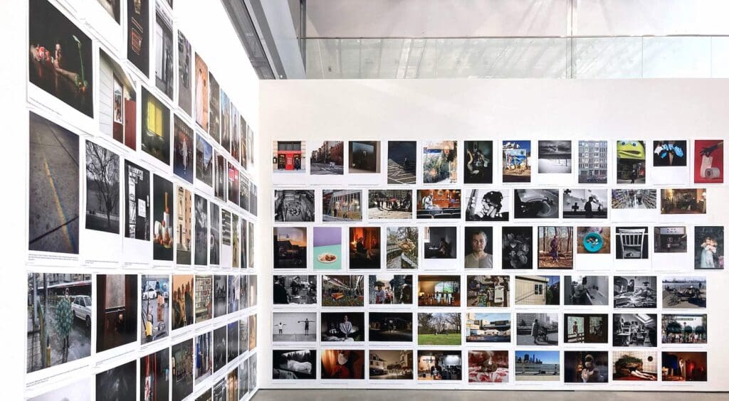 Art gallery exhibit with photographs arranged in a grid on two adjacent white walls in a modern space.