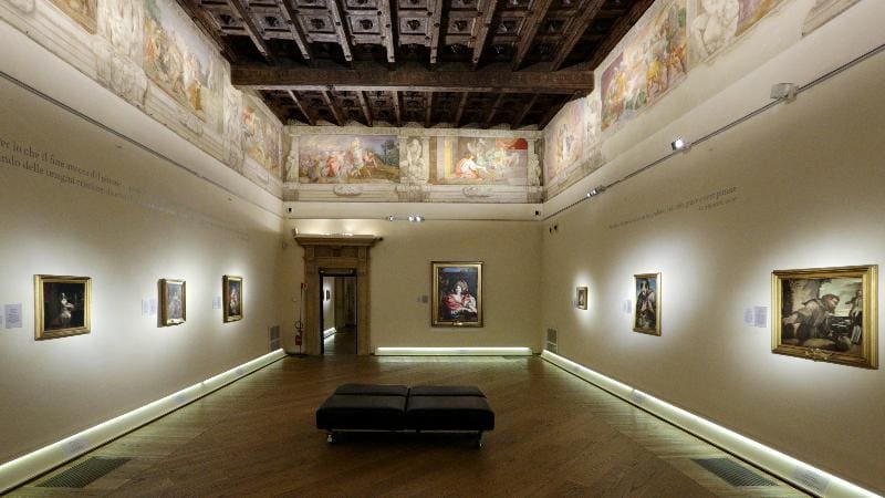 Art gallery with framed paintings, frescoed ceiling, modern lighting, and black cushioned bench.