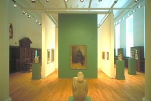 Art gallery interior with paintings and sculptures displayed on green and cream-colored walls.