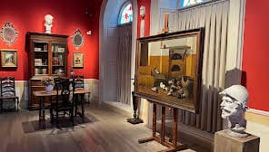 art gallery interior with red walls, painting on easel, bookshelves, bust, table and chairs