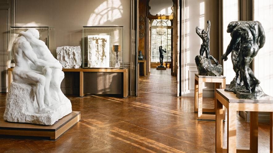 A well-lit gallery with polished floors displaying marble and bronze sculptures and adjacent gallery view.
