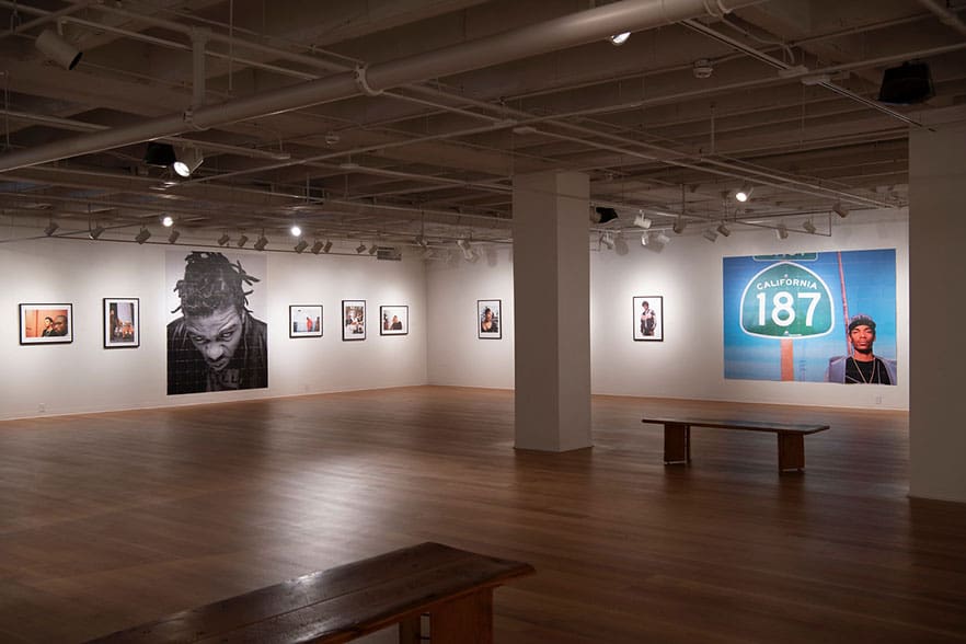 Art gallery with wooden floors, white walls, portraits, close-up and "California 187" sign, central bench.