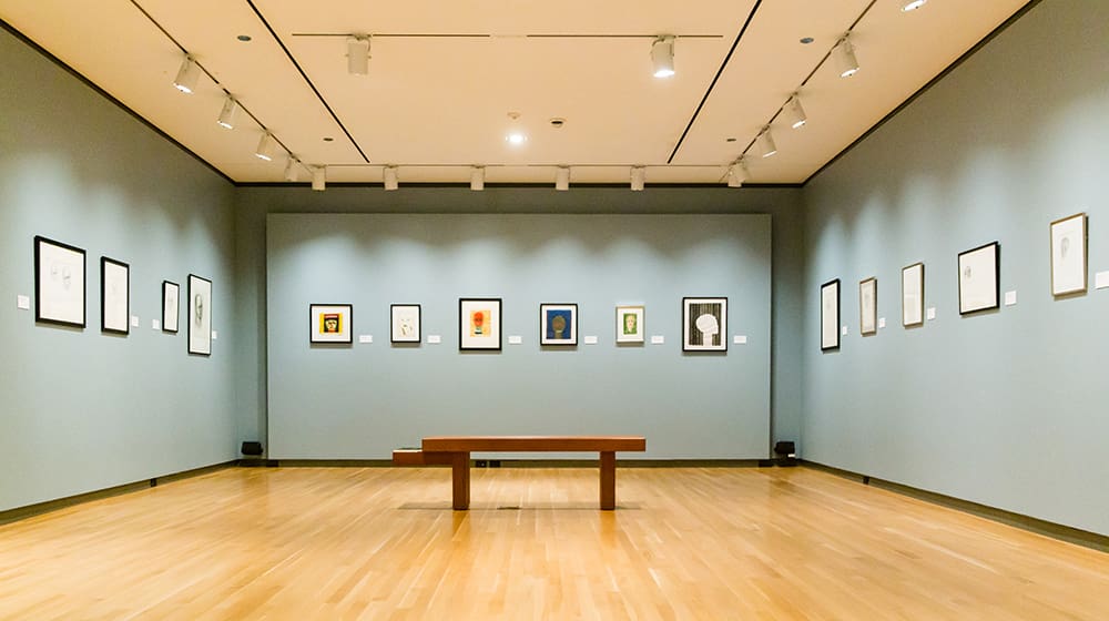 Well-lit art gallery with gray walls, wooden floor, framed artworks, and central wooden bench.