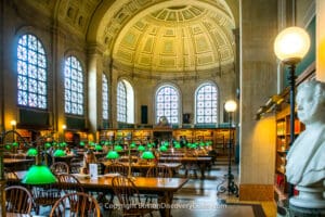 A bright library reading room with high ceilings, arched windows, green lamps, bookshelves, and a bust.