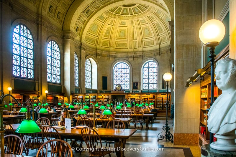A bright library reading room with high ceilings, arched windows, green lamps, bookshelves, and a bust.