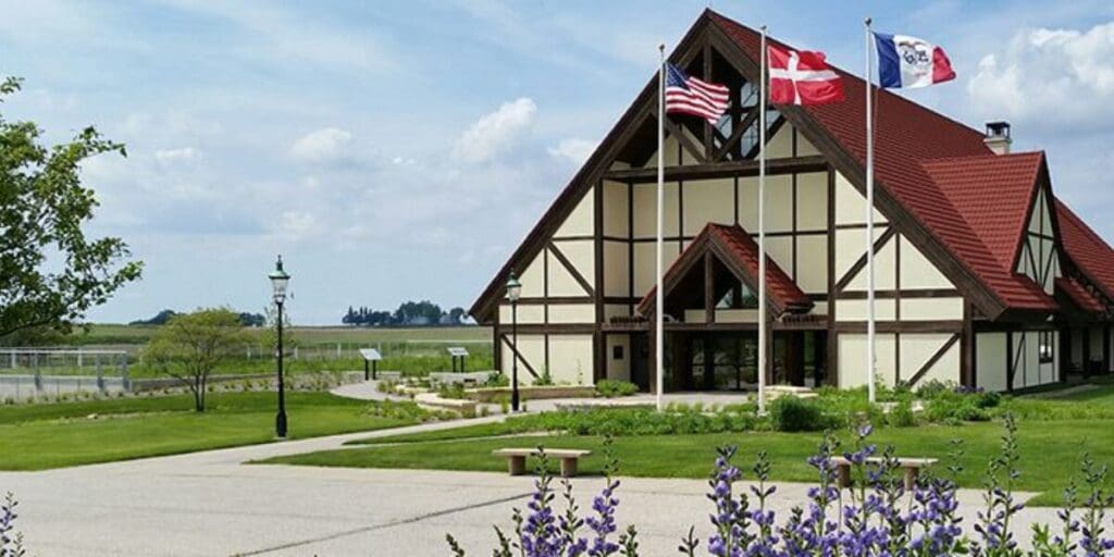 Building with steep red roof, timber-frame design, three flags, pathways, lawns, flowers, lamppost.