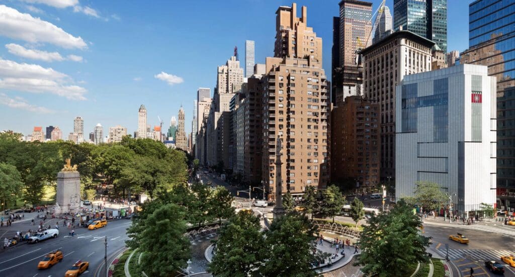Busy cityscape with a roundabout, fountain, trees, tall buildings, yellow taxis, pedestrians, blue sky