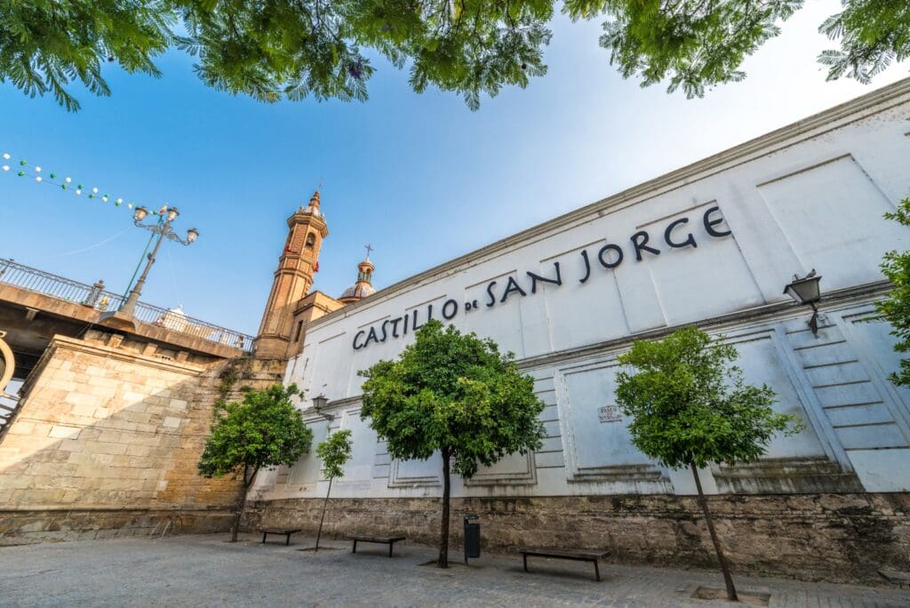 Entrance to Castillo de San Jorge with trees, adjacent structure, and tower under a clear blue sky