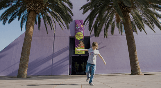 A child near a building with a purple facade, two palm trees, and a colorful banner at the entrance.