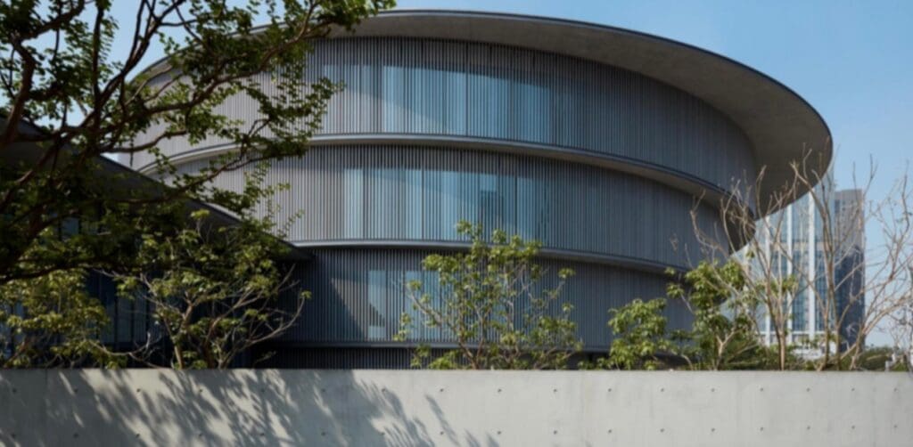 Circular multi-story building with horizontal slats and large windows, partially obscured by trees.