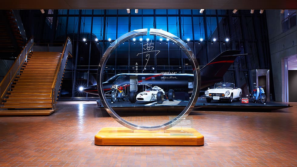 Circular metallic sculpture on a wooden base, with vehicles and large windows in a spacious showroom.