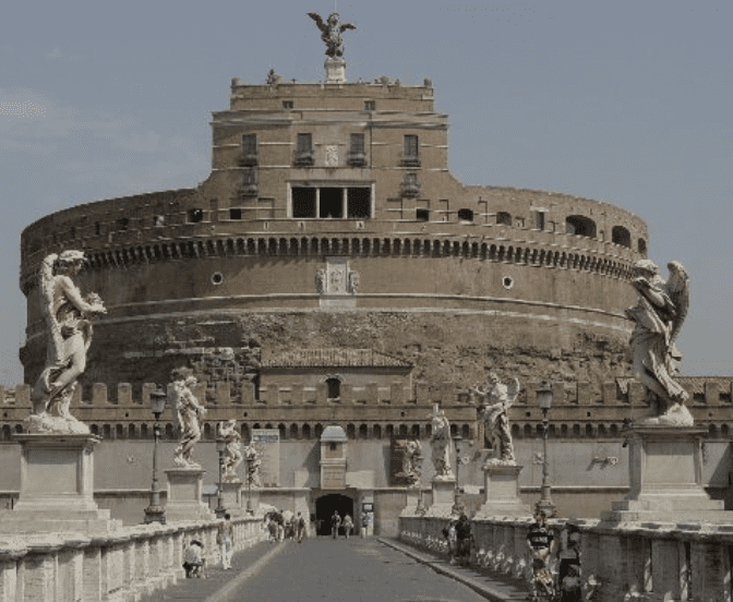Circular stone fortress with statues of angels along bridge, topped with angel statue, clear sky.