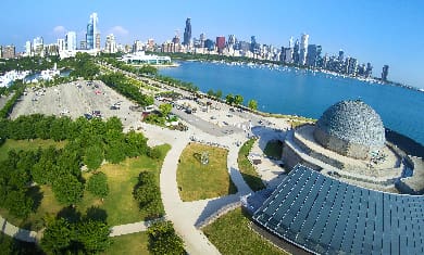 City skyline with waterfront, dome-shaped structure, paved walkway, green spaces under blue sky.