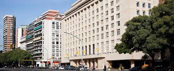 City street with parked vehicles, multi-story beige building, trees, and high-rise buildings.