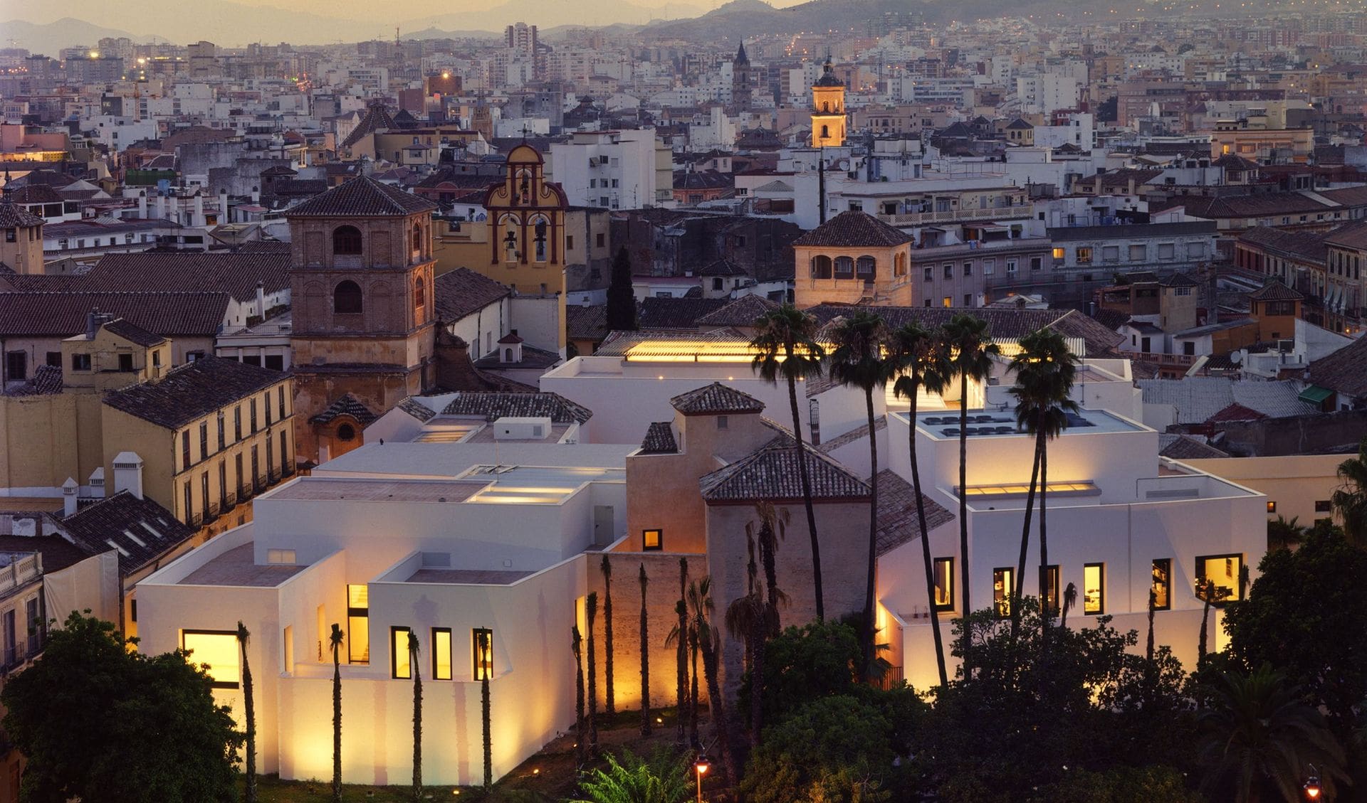 Cityscape at dusk with modern white building and historical structures, hills in the background.