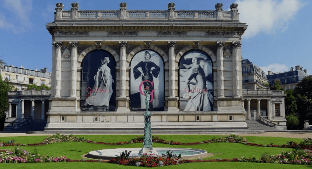 Classical building with fashion ads in arched windows, fountain and garden in foreground, blue sky.
