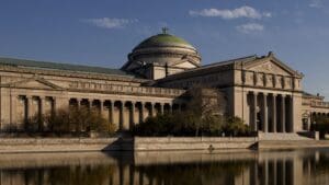 Classical-style building with a green dome, columns, next to a reflective body of water and trees.