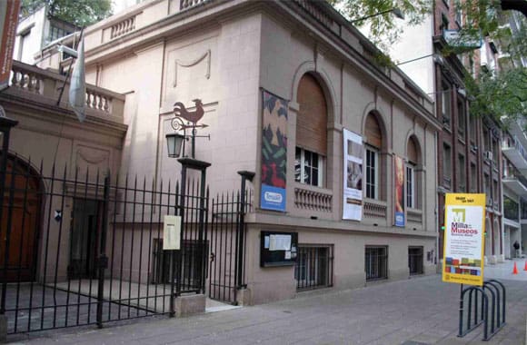 Classical building with arched windows, banners, wrought iron fence, and museum event sign on sidewalk.