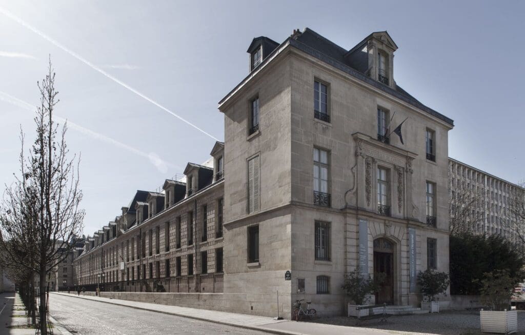 Large stone building with classical architecture, flag above entrance, cobblestone street with trees