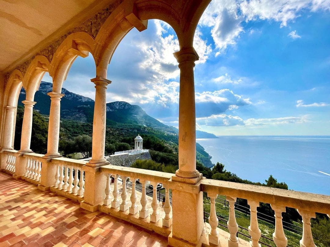 Coastal landscape viewed through arched columns with stone balustrades, hills, and blue waters.